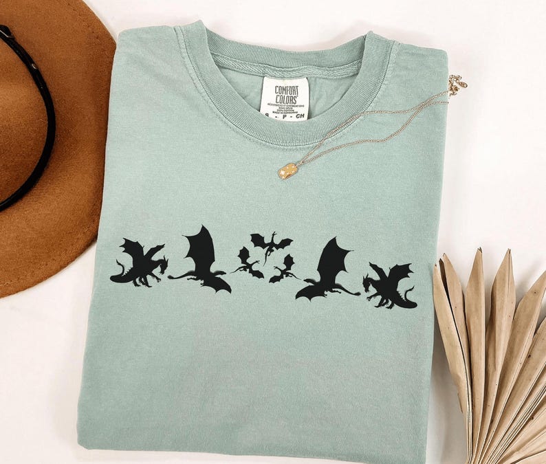 May include: A light green t-shirt with a black dragon graphic. The shirt has a crew neck and short sleeves. A gold necklace with a small pendant is on the shirt. A brown hat and dried plant are in the background.