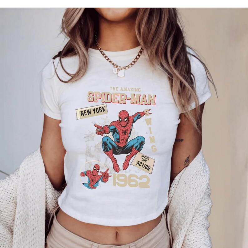 May include: Off-white t-shirt with a retro Spider-Man design. The print includes the words "The Amazing Spider-Man," "New York," and "Spring into Action," plus the year "1962." The graphic shows Spider-Man in his red and blue costume, in a dynamic pose.