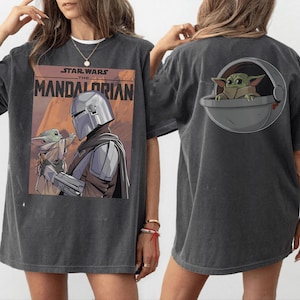 May include: Dark gray t-shirt featuring a graphic from "Star Wars: The Mandalorian." The front displays the show's title and an image of the Mandalorian and Grogu. The back has a Grogu graphic in a pod.
