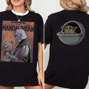 May include: Black t-shirt featuring a graphic from the Star Wars series, "The Mandalorian." The front of the shirt displays the Mandalorian and Grogu. The back of the shirt shows Grogu in a pod. The shirt has a white collar.