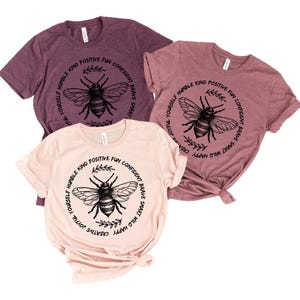 May include: Three short-sleeved t-shirts in shades of mauve and blush. Each shirt features a circular design with a bee illustration and the words "Yourself, Humble, Kind, Positive, Fun, Confident, Brave, Smart, Wild, Happy, Creative, Joyful".