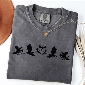 May include: A gray Comfort Colors t-shirt featuring a black dragon graphic. The shirt has a crew neck and short sleeves. A gold necklace with a pendant is draped across the shirt. A brown hat and dried leaves are in the background.