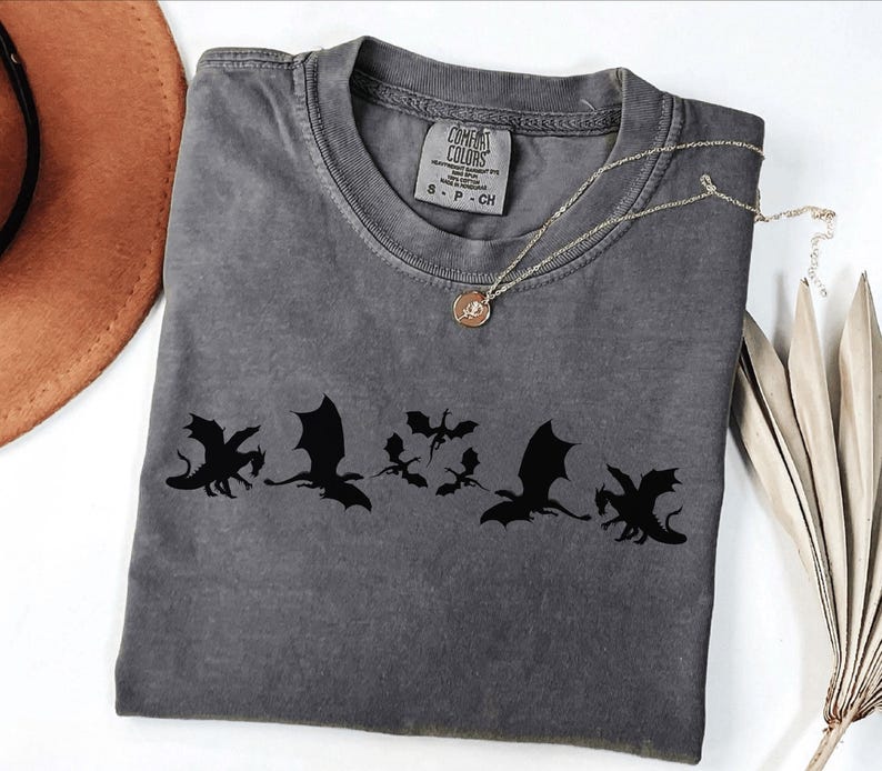 May include: A gray Comfort Colors t-shirt featuring a black dragon graphic. The shirt has a crew neck and short sleeves. A gold necklace with a pendant is draped across the shirt. A brown hat and dried leaves are in the background.