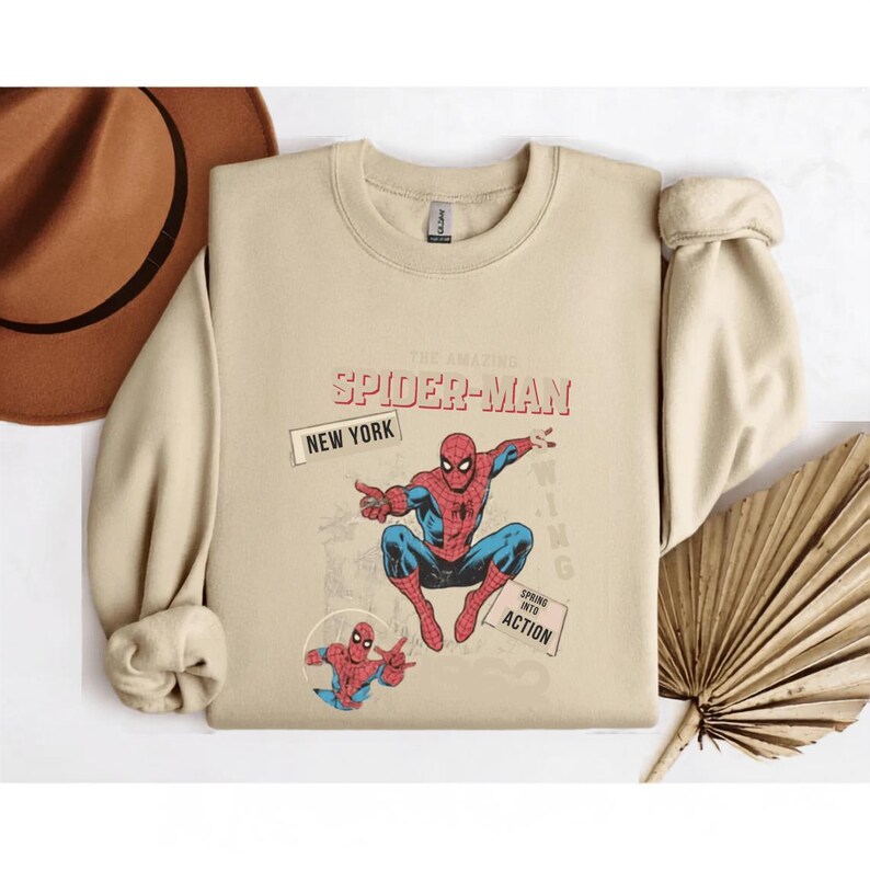 May include: Beige crew neck sweatshirt with a Spider-Man graphic. The design includes the text "THE AMAZING SPIDER-MAN" and "NEW YORK". A smaller Spider-Man graphic and the words "SPRING INTO ACTION" are also present. The sweatshirt has rolled cuffs.