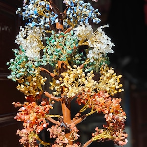 May include: A decorative tree sculpture with a brown trunk and branches. The tree's foliage is made of various colourful gemstones, including blue, green, yellow, orange, and clear crystals, attached with gold wire. A unique home decor item.