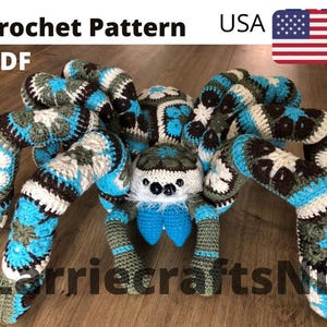 Puede incluir: Una araña grande de ganchillo en tonos azules, marrones, blancos y verdes. La araña tiene ocho patas largas y segmentadas y un cuerpo redondo. El texto "Crochet Pattern PDF USA" está en la parte superior. Es una artesanía hecha a mano.