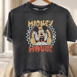 Peut inclure: Un t-shirt gris anthracite avec un motif Mickey Mouse de style vintage. Le design comprend les mots "Mickey Mouse" dans une police r&eacute;tro, avec Mickey au centre. Le t-shirt est suspendu &agrave; un cintre en bois.