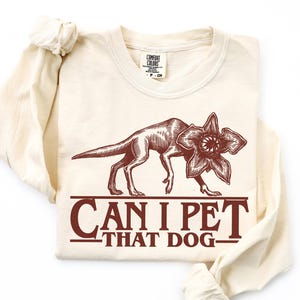 May include: Cream-colored long-sleeve shirt featuring a Demogorgon illustration in brown. The text "CAN I PET THAT DOG" is printed below the creature. The shirt is made of a soft material.