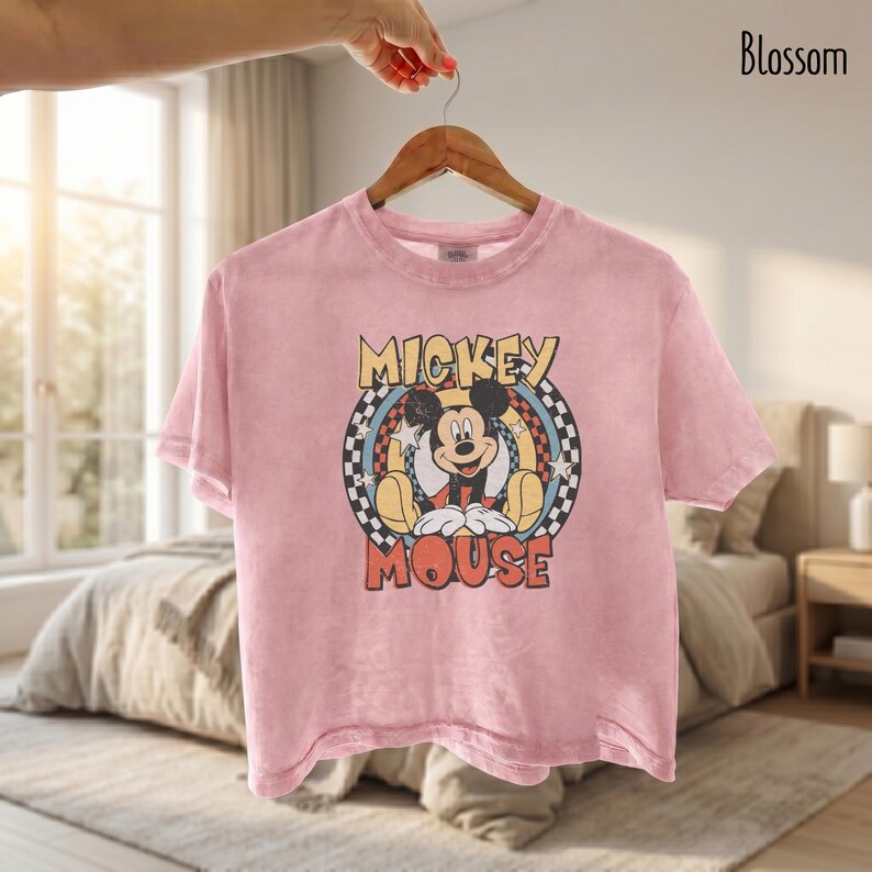Peut inclure: T-shirt rose clair avec un motif de Mickey Mouse. Le design comprend les mots "Mickey Mouse" dans une police r&eacute;tro, avec Mickey au centre. Le t-shirt est suspendu &agrave; un cintre en bois.