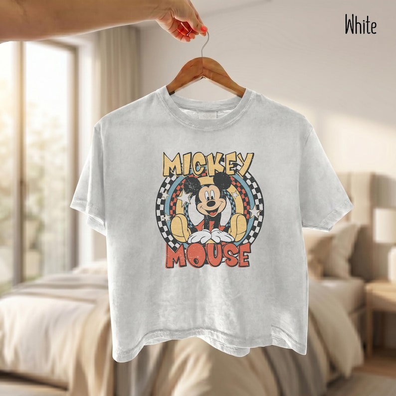 Peut inclure: T-shirt blanc &agrave; manches courtes avec un motif de Mickey Mouse. Le design comprend les mots "Mickey Mouse" dans une police r&eacute;tro, sur un fond &agrave; carreaux et &agrave; &eacute;toiles. Le t-shirt est suspendu &agrave; un cintre en bois.