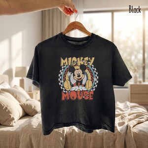 Peut inclure: T-shirt noir avec un motif vintage de Mickey Mouse. Le design comprend les mots "Mickey Mouse" dans une police r&eacute;tro, avec Mickey au centre. Le t-shirt est suspendu &agrave; un cintre en bois.