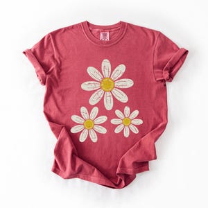 May include: A red t-shirt featuring a daisy graphic. The graphic includes three daisies with white petals and yellow centers. The shirt has a crew neck and short sleeves, with the sleeves rolled up.
