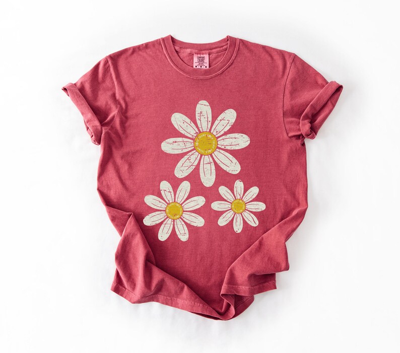 May include: A red t-shirt featuring a daisy graphic. The graphic includes three daisies with white petals and yellow centers. The shirt has a crew neck and short sleeves, with the sleeves rolled up.
