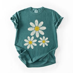 May include: Teal-colored t-shirt featuring a daisy graphic. The graphic includes three white daisies with yellow centers. The shirt has a classic crew neck and short sleeves, perfect for casual wear.