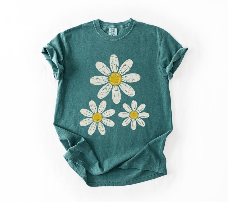 May include: Teal-colored t-shirt featuring a daisy graphic. The graphic includes three white daisies with yellow centers. The shirt has a classic crew neck and short sleeves, perfect for casual wear.