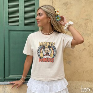 Peut inclure: T-shirt couleur ivoire avec un motif vintage de Mickey Mouse. Le motif comprend Mickey Mouse dans une pose assise dans un motif &agrave; carreaux circulaire, avec les mots "Mickey Mouse". Le t-shirt a des manches courtes.