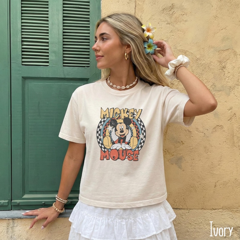 Peut inclure: T-shirt couleur ivoire avec un motif vintage de Mickey Mouse. Le motif comprend Mickey Mouse dans une pose assise dans un motif &agrave; carreaux circulaire, avec les mots "Mickey Mouse". Le t-shirt a des manches courtes.