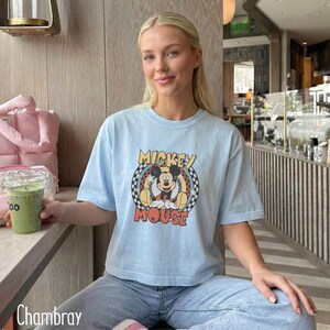 Peut inclure: T-shirt bleu clair avec un motif de Mickey Mouse dans un cercle &agrave; carreaux et les mots "Mickey Mouse". Le t-shirt a des manches courtes et est fait d'une mati&egrave;re douce. Le mot "Chambray" est en bas &agrave; gauche.