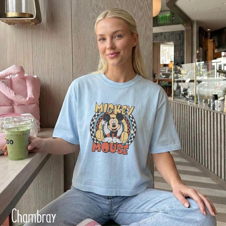 Peut inclure: T-shirt bleu clair avec un motif de Mickey Mouse dans un cercle &agrave; carreaux et les mots "Mickey Mouse". Le t-shirt a des manches courtes et est fait d'une mati&egrave;re douce. Le mot "Chambray" est en bas &agrave; gauche.