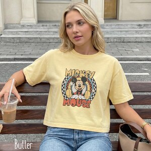 Peut inclure: T-shirt jaune beurre avec un motif Mickey Mouse de style vintage. Le motif comprend les mots "Mickey Mouse" dans une police r&eacute;tro, sur un fond &agrave; carreaux et &eacute;toil&eacute;. Le t-shirt est port&eacute; avec un jean bleu.