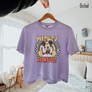 Peut inclure: Un t-shirt violet avec un motif de Mickey Mouse. Le design comprend les mots "Mickey Mouse" en jaune et rouge, avec Mickey au centre. Le t-shirt est suspendu &agrave; un cintre en bois.