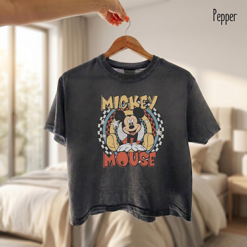 Peut inclure: Un t-shirt gris anthracite avec un motif Mickey Mouse de style vintage. Le design comprend les mots "Mickey Mouse" dans une police r&eacute;tro, avec Mickey au centre. Le t-shirt est suspendu &agrave; un cintre en bois.