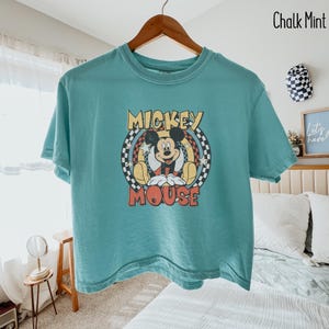Peut inclure: Un t-shirt de couleur sarcelle avec un motif de Mickey Mouse. Le design comprend les mots "Mickey Mouse" dans une police vintage, avec Mickey au centre. Le t-shirt est suspendu &agrave; un cintre en bois.