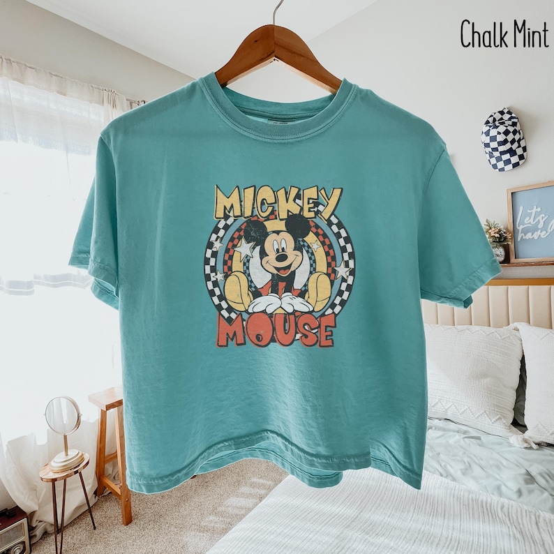 Peut inclure: Un t-shirt de couleur sarcelle avec un motif de Mickey Mouse. Le design comprend les mots "Mickey Mouse" dans une police vintage, avec Mickey au centre. Le t-shirt est suspendu &agrave; un cintre en bois.