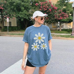 May include: A blue t-shirt with three daisy graphics. The daisies have white petals and yellow centers. The shirt is a relaxed fit and has rolled-up sleeves. The person is wearing a white cap and sunglasses.