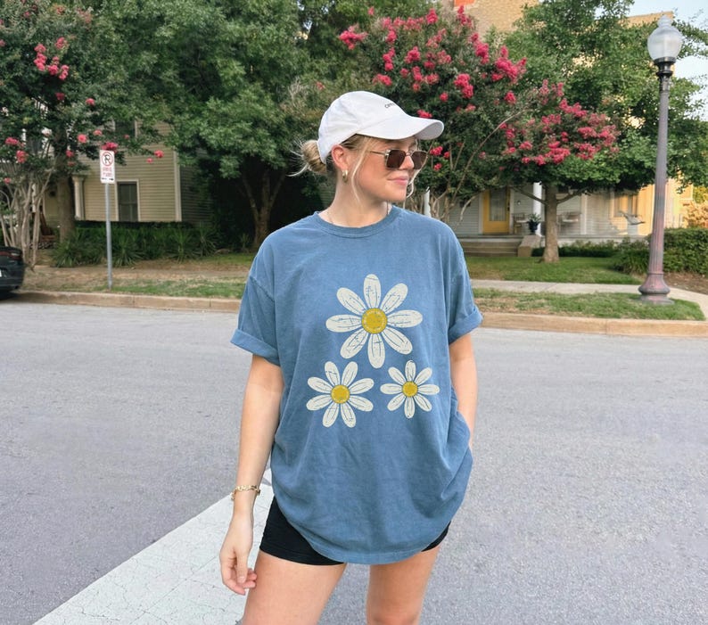 May include: A blue t-shirt with three daisy graphics. The daisies have white petals and yellow centers. The shirt is a relaxed fit and has rolled-up sleeves. The person is wearing a white cap and sunglasses.