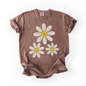 May include: A brown t-shirt featuring a daisy graphic. The graphic has three daisies with white petals and yellow centers. The t-shirt is folded, showing the rolled sleeves and the tag at the neckline.