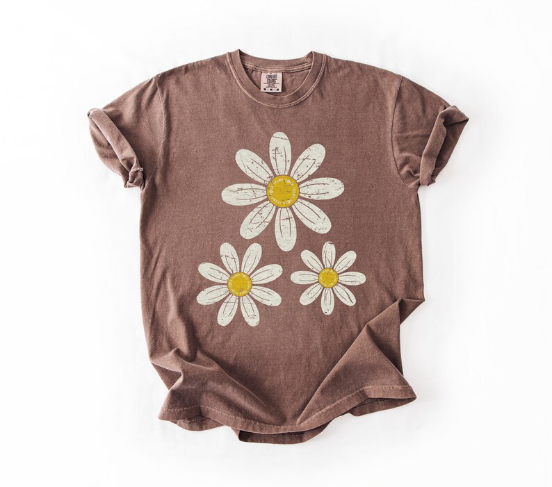 May include: A brown t-shirt featuring a daisy graphic. The graphic has three daisies with white petals and yellow centers. The t-shirt is folded, showing the rolled sleeves and the tag at the neckline.