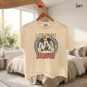 Peut inclure: T-shirt ivoire avec un motif de Mickey Mouse. Le design comprend les mots "Mickey Mouse" dans une police r&eacute;tro, avec Mickey au centre. Le t-shirt est suspendu &agrave; un cintre en bois.