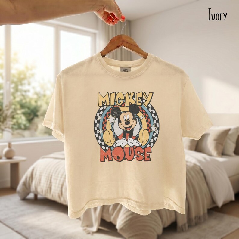 Peut inclure: T-shirt ivoire avec un motif de Mickey Mouse. Le design comprend les mots "Mickey Mouse" dans une police r&eacute;tro, avec Mickey au centre. Le t-shirt est suspendu &agrave; un cintre en bois.
