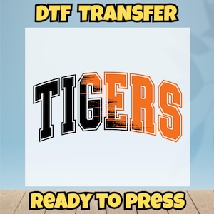 May include: A DTF transfer design featuring the word "TIGERS" in black and orange block letters. The text is arched across a white background. The words "DTF TRANSFER" and "READY TO PRESS" are in yellow block letters.