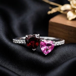 May include: A silver ring featuring two heart-shaped gemstones, one deep red and one bright pink, set side-by-side. The band is crafted from silver and is encrusted with small, clear, sparkling stones along the shank, creating a refined look.