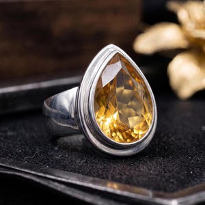 May include: A silver ring featuring a large, faceted, pear-shaped yellow citrine gemstone set in a smooth metal bezel. The band is polished silver, and the stone displays a warm, golden-yellow hue with multiple triangular facets reflecting light.