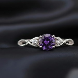 May include: A silver ring with a large, round, purple gemstone at the center, flanked by two smaller clear stones. The band features a twisted design. The ring is set against a dark, textured background.