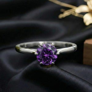 May include: A silver ring with a large, round, deep purple gemstone. The ring is set on a black, textured surface. The gemstone is the focal point, catching the light and showcasing its facets.