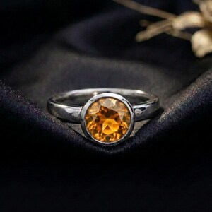 May include: A silver ring featuring a round, faceted, golden-yellow citrine gemstone set in a smooth bezel setting. The band of the ring is polished and has a simple, clean design that highlights the vibrant color and clarity of the central stone.