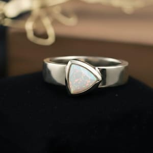 Könnte beinhalten: Ein silberner Ring mit einem dreieckigen, weißen Opal-Edelstein. Der Opal hat ein Farbspiel mit roten und orangen Blitzen. Der Ring ist breit und poliert, was einen modernen und eleganten Look erzeugt.