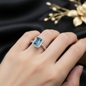 May include: A silver ring featuring a rectangular, light blue gemstone surrounded by a halo of smaller, clear stones. The ring is set against a dark background, highlighting the sparkle of the gems. A classic piece of jewelry.
