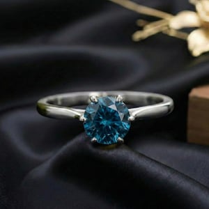 May include: A silver ring with a large, round, deep blue gemstone. The ring is set against a black satin background, with the gemstone prominently displayed. The ring's band is smooth and polished.