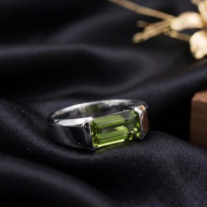 May include: A silver-toned metal ring featuring a rectangular, emerald-cut, lime-green gemstone set horizontally in the center. The band is smooth and polished, showcasing a clean, metallic finish that reflects light across its surface.