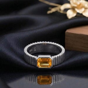 May include: A silver ring with a rectangular, yellow gemstone. The ring has a ribbed band and a polished finish. The gemstone is set in a bezel setting. The ring is displayed on a black satin surface.
