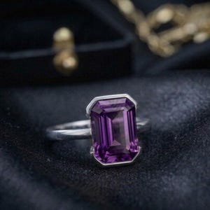 May include: A silver ring with a large, rectangular, deep purple gemstone. The gemstone is set in a silver bezel, and the ring is displayed on a dark, textured surface. The ring is a piece of jewelry.