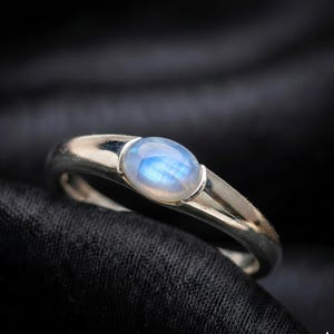 May include: A silver ring with an oval, iridescent, blue-white gemstone. The ring's band is smooth and polished, with a split design near the gemstone. The gemstone is set in a bezel setting, and the ring is displayed against a dark background.