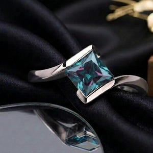 May include: A silver ring with a square, teal and purple gemstone. The gemstone is set in a silver bezel. The ring is displayed on a black, textured surface. The ring's band curves gracefully.