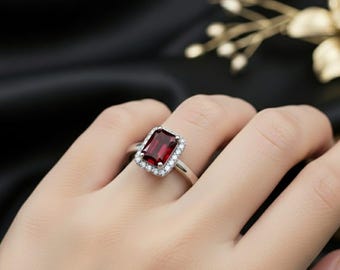 May include: A silver ring featuring a rectangular, deep red gemstone surrounded by a halo of smaller, clear stones. The ring is set on a silver band and is displayed on a hand against a dark background.