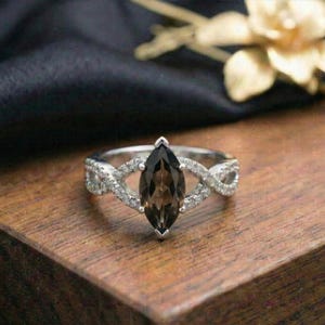 May include: A silver ring with a marquise-cut, smoky brown gemstone as the focal point. The band features an infinity design with small, clear accent stones. The ring is set on a wooden surface with a dark background.